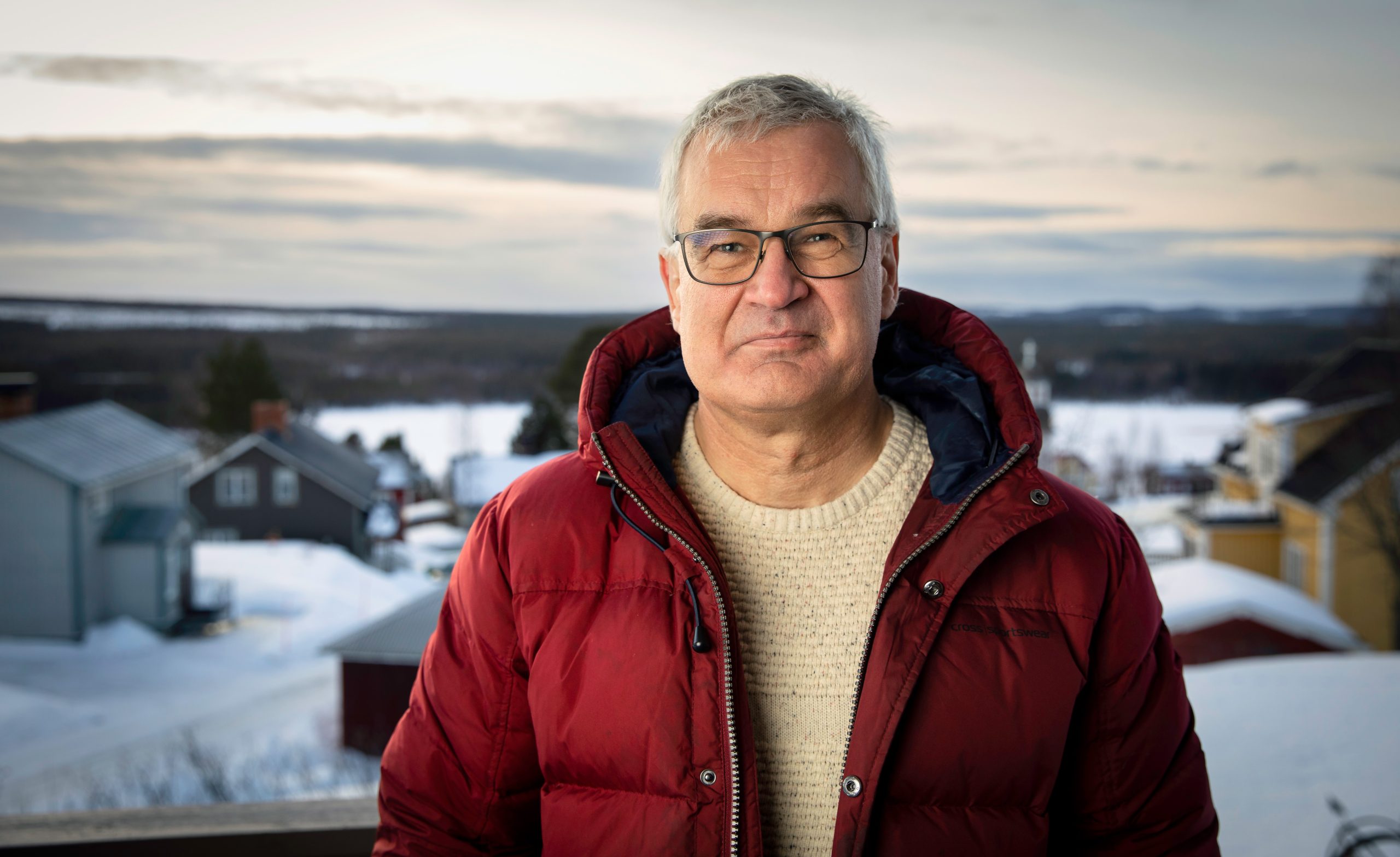 Lars Blixt tittar in i kameran i röd jacka framför en suddig bakgrund av hus och snö